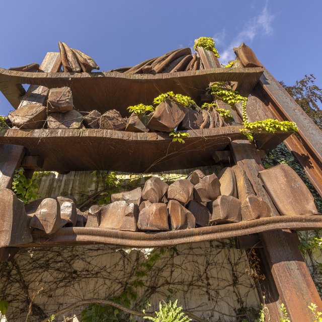 Rostige Stahlskulptur mit aufgeschichteten Metall- und Steinformen, überwachsen von Pflanzen, im öffentlichen Raum in Peine.