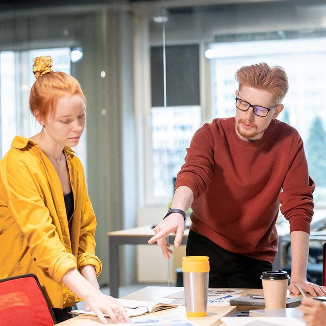 Drei junge Personen stehen um einen Tisch mit Papieren und Kaffeebechern in einem modernen Büro und diskutieren.