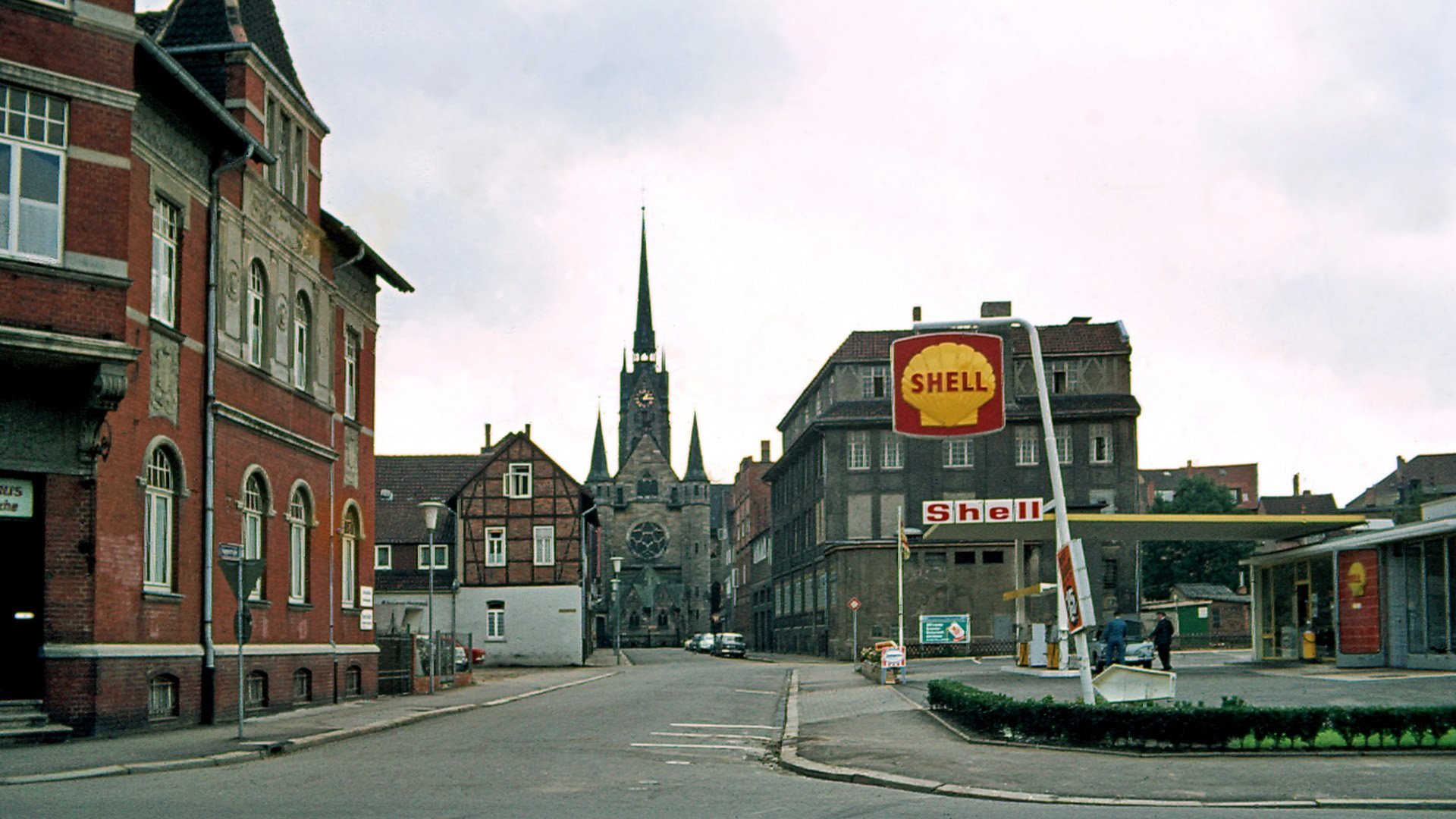 Eine Straße mit alten Backsteinhäusern und einer Shell-Tankstelle, im Hintergrund eine Kirche.