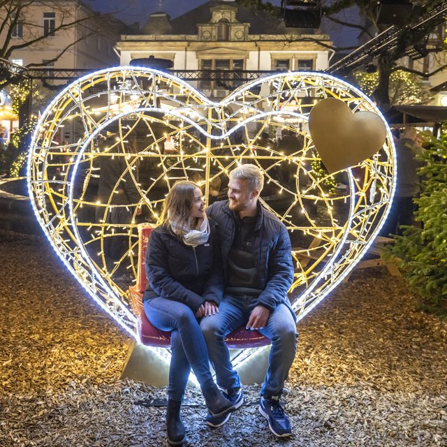 Paar sitzt in einer beleuchteten Herz-Lichtskulptur auf der Weihnachtsstadt in der Innenstadt von Peine, umgeben von geschmückten Tannen und warmem Lichterglanz am Abend.