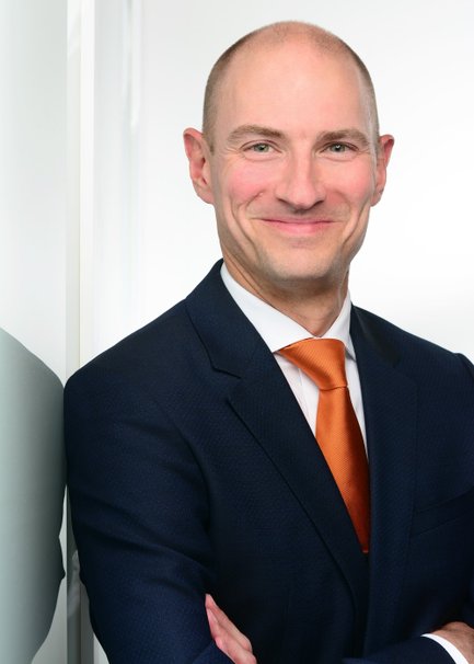 Porträt von einem Mann im dunklen Anzug mit orangefarbener Krawatte vor heller Wand.