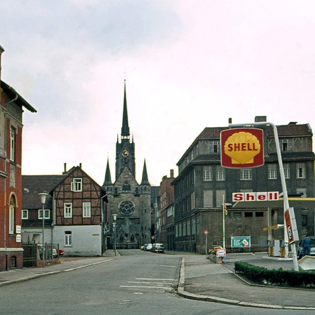 Eine Straße mit alten Backsteinhäusern und einer Shell-Tankstelle, im Hintergrund eine Kirche.