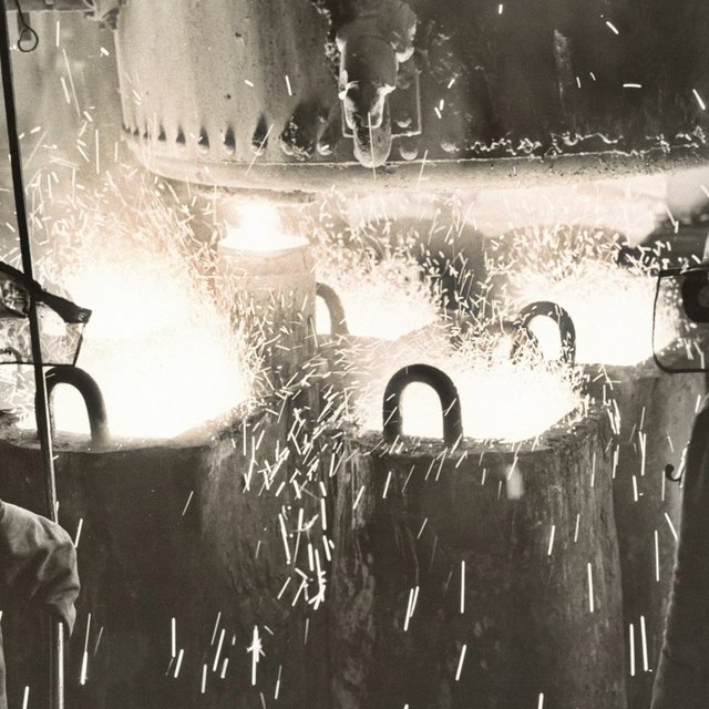 Historische Schwarz-Weiß-Aufnahme aus dem Peiner Stahlwerk. Mehrere Arbeiter stehen in einer Werkhalle beim Kokillenguss. Im Hintergrund leuchtet das flüssige, heiße Metall hell auf.