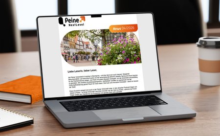 Laptop auf einem Schreibtisch mit geöffneter digitalen Ausgabe des Newsletters „Peine NextLevel News 04/2026“. Auf dem Bildschirm ist ein Beitrag zur Entwicklung der Peiner Innenstadt mit einem Foto der Innenstadt und blühenden Blumen zu sehen. Neben dem Laptop stehen ein Coffee-to-go-Becher, ein orangefarbenes Notizbuch und ein Schreibblock.