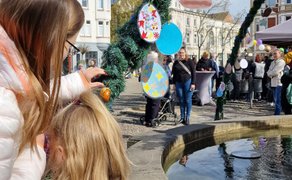 Kind schmückt gemeinsam mit einer Begleitperson den Osterbrunnen auf dem Marktplatz in Peine mit bunten Ostereiern.