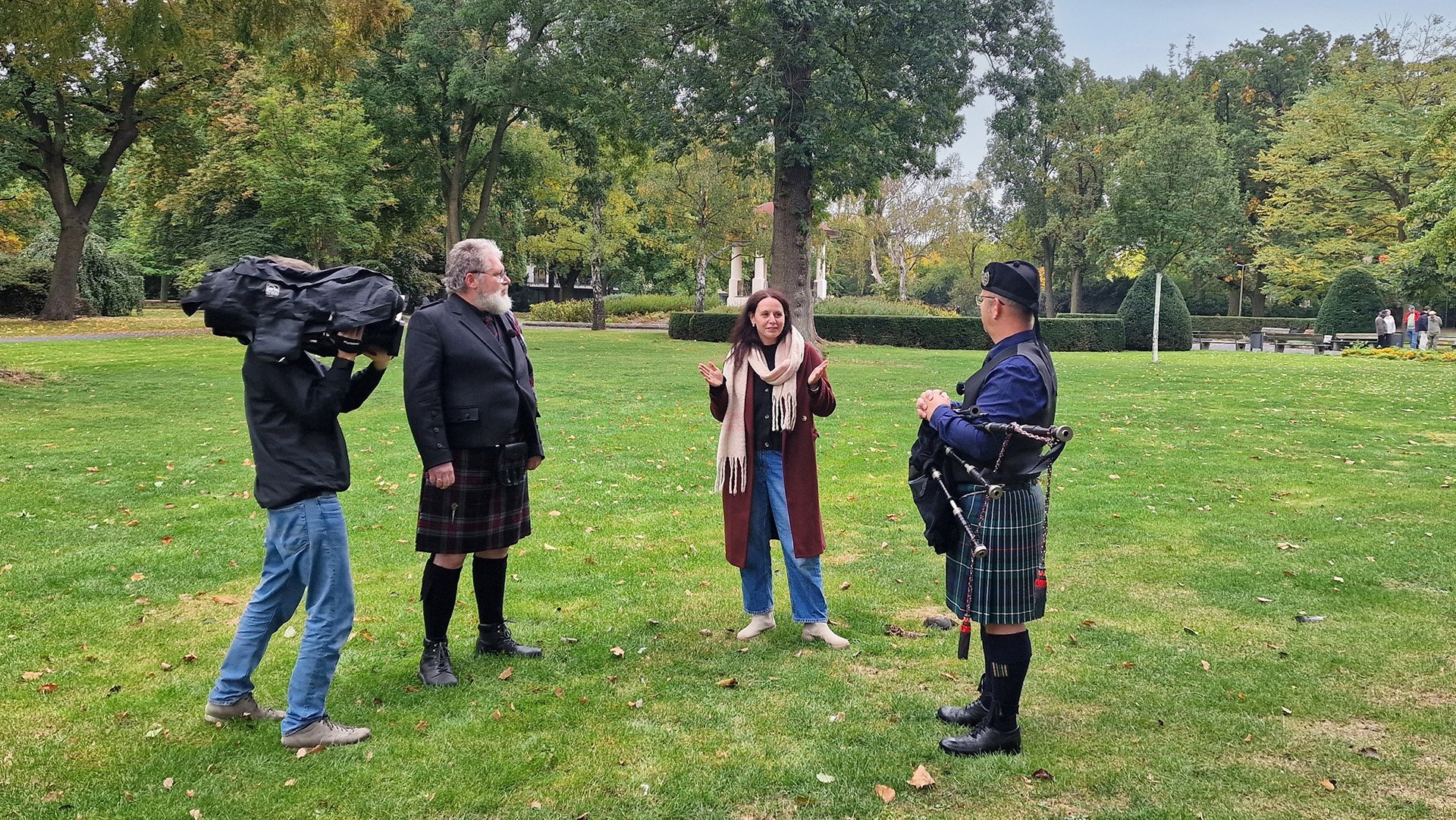 Ein Mann mit Kamera filmt zwei Männer im Kilt und eine Frau in einem Park.