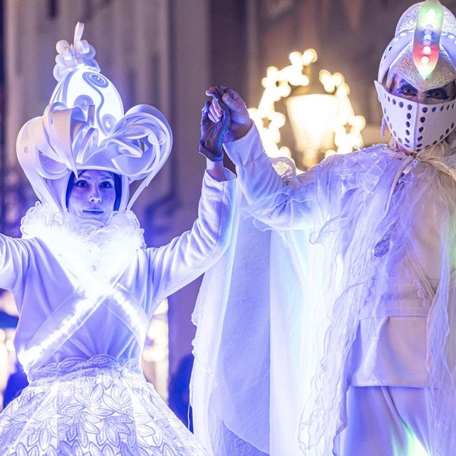 Zwei Personen in leuchtenden, weißen Kostümen mit Lichtern, eine hält eine leuchtende Kugel, die andere trägt einen leuchtenden Kopfschmuck.