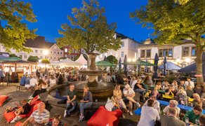 Peiner Weinfest am Abend auf dem Marktplatz mit beleuchtetem Brunnen, vielen Besucherinnen und Besuchern, roten Sitzsäcken, grünen Sonnenschirmen, Weinständen und sommerlicher Atmosphäre in der Innenstadt von Peine.