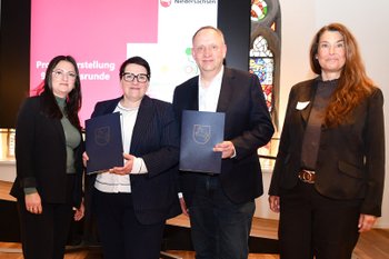 Vier Personen stehen bei der Übergabe von Förderbescheiden des Programms „Zukunftsräume Niedersachsen“ vor einem Bildschirm mit Präsentationsfolie in einem historischen Saal.