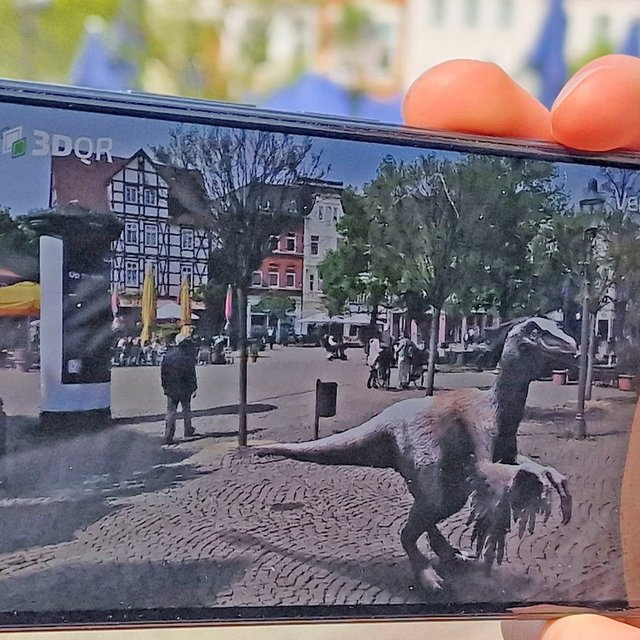 Augmented-Reality-Ansicht eines Dinosauriers, der über ein Smartphone in einen belebten Stadtplatz eingeblendet wird. Auf dem Display ist die virtuelle Figur in realistischer Größe zwischen Passanten, Bäumen und historischen Gebäuden zu sehen, während die reale Umgebung im Hintergrund erkennbar bleibt.