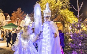 Zwei Personen in weißen, leuchtenden Kostümen mit Kronen und Zepter auf einem festlich beleuchteten Weihnachtsmarkt im Freien bei Nacht.
