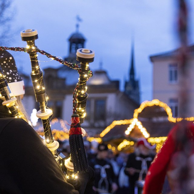 Spieler einer Dudelsackgruppe mit beleuchteten Instrumenten bei einem Weihnachtsmarkt vor historischen Gebäuden in der Dämmerung.