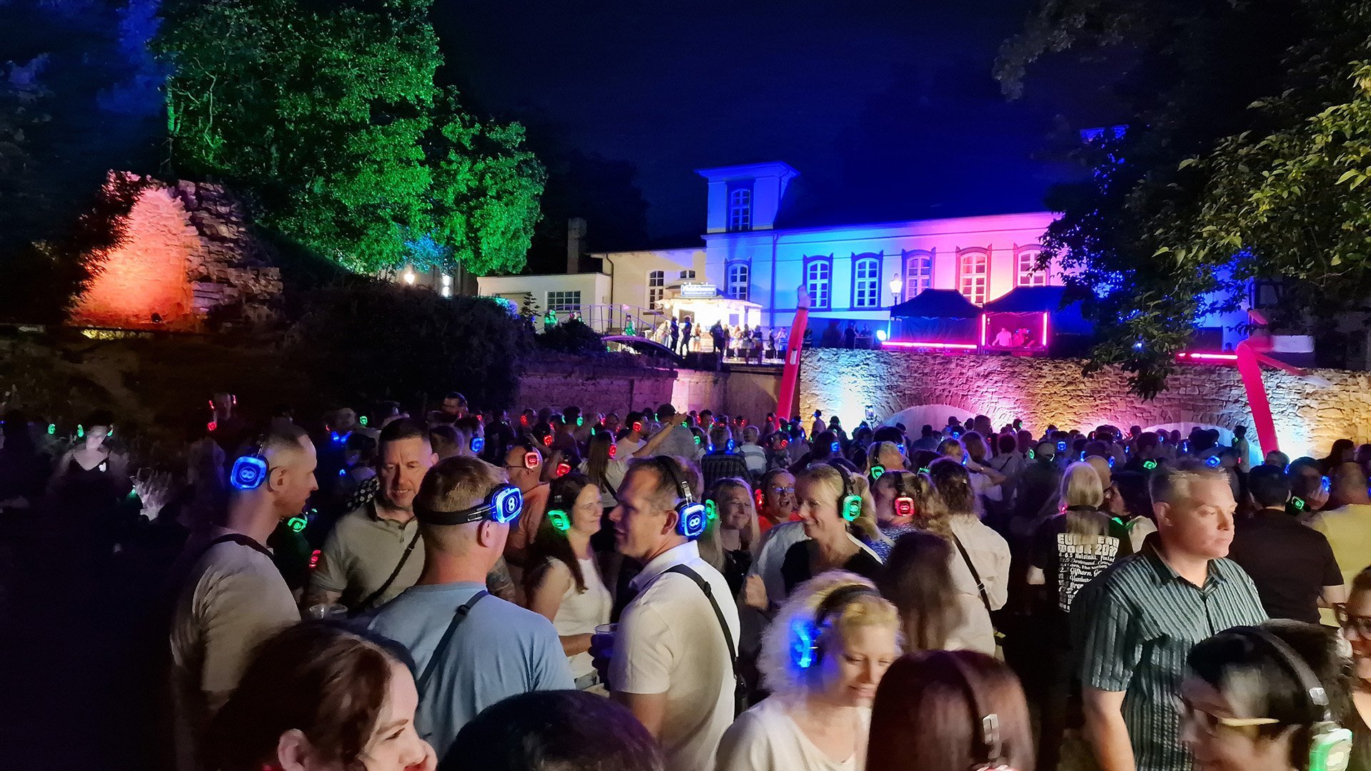 Große Menschenmenge mit leuchtenden Kopfhörern bei einer Open-Air-Party vor einem beleuchteten Gebäude in der Nacht.