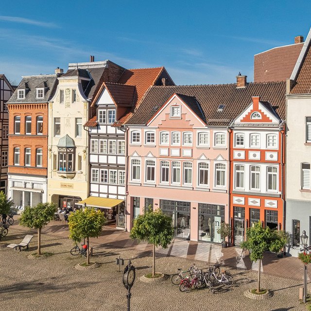 Historische Fachwerkhäuser und farbenfrohe Gebäude säumen einen sonnigen Marktplatz mit Bäumen, Sitzgelegenheiten und Fahrrädern in der Peiner Innenstadt.