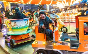 Kind sitzt auf orangefarbenem Karussellauto, umgeben von bunten, beleuchteten Fahrgeschäften auf Weihnachtsmarkt.