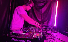 DJ mit Kopfhörern steht an einem beleuchteten Mischpult in pink-violettem Licht und legt Musik in einer dunklen Festival-Atmosphäre auf.