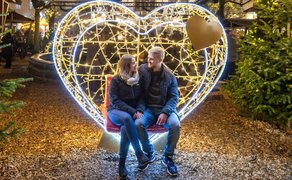 Zwei Personen sitzen auf einer roten Bank vor einem großen, herzförmigen Lichterbogen mit goldfarbenem Herz an der Seite, umgeben von Tannenbäumen.