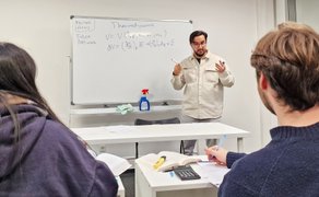 Ein Mann steht vor einem Whiteboard mit Formeln und spricht in Richtung von zwei Personen, die sich Notizen machen.
