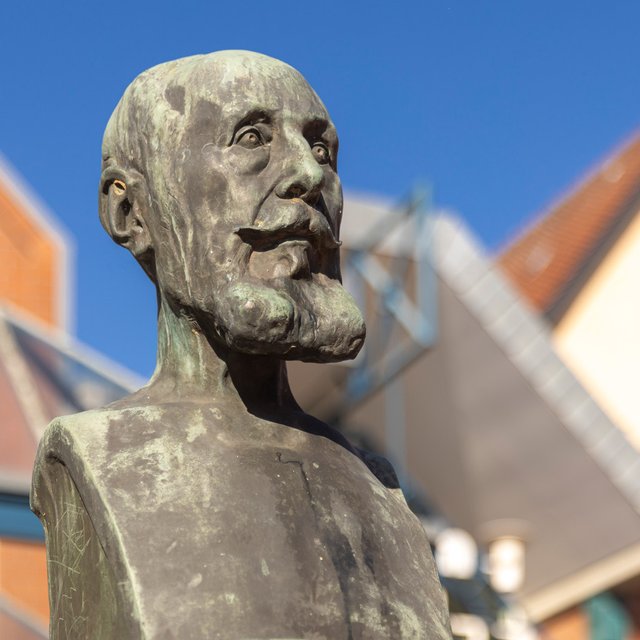 Nahaufnahme einer bronzenen Statue eines Mannes mit markantem Bart und ernstem Gesichtsausdruck. Die Figur ist detailreich gearbeitet und blickt leicht nach oben. Im Hintergrund sind unscharf Gebäude mit roten Ziegeldächern und ein klarer blauer Himmel zu sehen. Die Szene zeigt ein Denkmal im städtischen Raum bei sonnigem Wetter.