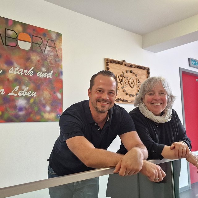 Ein Mann und eine Frau lehnen an einem Geländer, dahinter eine bunte Wandtafel mit der Aufschrift „LABORA bunt, stark und voller Leben“.