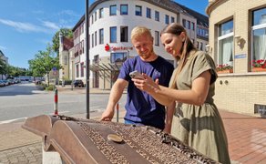 Zwei Teilnehmende nutzen ein Smartphone und eine Infostation in der Innenstadt von Peine während einer digitalen Stadtrallye.“