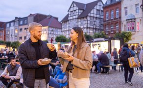 Ein Mann und eine Frau stehen mit Getränken auf einem belebten Platz, im Hintergrund sitzen und stehen weitere Menschen vor Fachwerkhäusern.