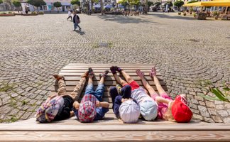 Das Bild zeigt vier Kinder, die auf eine Holzliege auf dem Historischen Marktplatz in Peine liegen. 