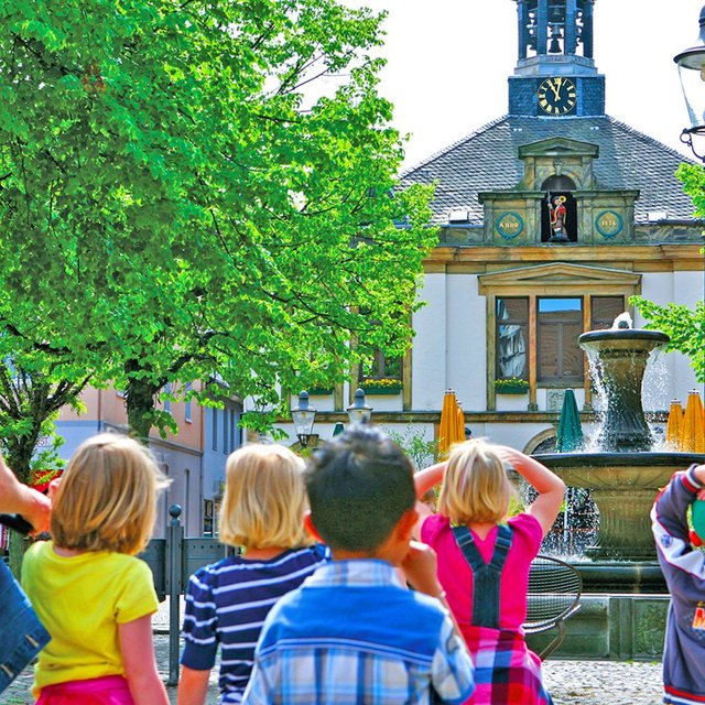 Eine Gruppe von Kindern steht gemeinsam mit einer erwachsenen Begleitperson auf einem Platz und blickt in Richtung eines historischen Gebäudes mit Uhrturm. Im Vordergrund sprudelt ein Brunnen, umgeben von Bäumen und Sitzgelegenheiten. Die Kinder schauen interessiert nach vorne und folgen aufmerksam einer Stadtführung in der lebendigen Innenstadt.