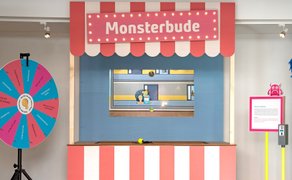 Wurfbude mit rot-weiß gestreiftem Baldachin und Schild „Monsterbude“, links ein Glücksrad, rechts ein Infotext und Monster-Figuren auf einem Ständer.