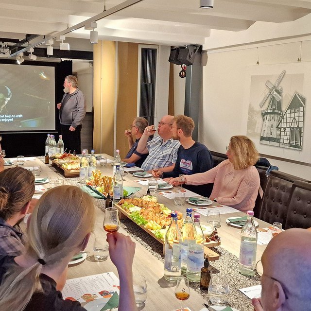 Eine Gruppe von Menschen sitzt gemeinsam an einem Tisch mit Käseplatte und Gläsern und verfolgt eine Präsentation.