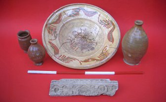 Vier archäologische Keramikgefäße und ein verziertes Steinfragment auf rotem Untergrund.