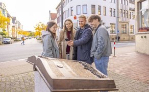 Vier junge Erwachsene lösen gemeinsam ein Rätsel mit Smartphone an einer Infotafel bei der PeineCode-Stadtrallye in der Peiner Innenstadt.