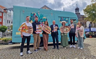 Sechs Personen stehen nebeneinander auf einem Platz vor einem türkisfarbenen Foodtruck.