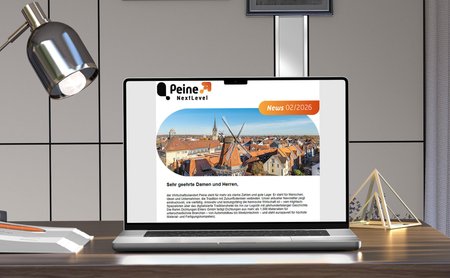 Laptop auf einem Schreibtisch zeigt den Newsletter „Peine NextLevel – News 02/2026“ mit Luftaufnahme der Peiner Innenstadt und Windmühle auf dem Bildschirm.