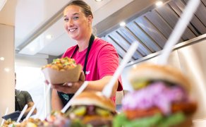 Eine Frau hinter einer Theke hält eine Schale mit Essen in den Händen, im Vordergrund sind Speisen zu sehen.