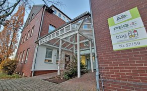 Eingangsbereich eines roten Backsteingebäudes mit Glasvordach und Schild mit den Logos von A+P, PEG und bbg. 