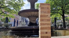 Hoher Turm aus gestapelten Holzklötzen steht am Rand eines steinernen Brunnens; im Hintergrund sprudelt Wasser, umgeben von Bäumen, Sonnenschirmen und Gebäuden auf einem belebten Marktplatz.