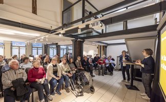 Eine Gruppe von rund 30 überwiegend älteren Menschen sitzt in einem modernisierten Fachwerkraum und hört aufmerksam einer Referentin zu, die rechts neben einer Präsentationsfläche steht und einen Vortrag hält.