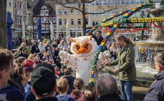 Das Maskottchen Uhlinchen steht auf dem Peiner Marktplatz neben dem geschmückten Osterbrunnen und begrüßt Kinder und Besucher, während viele Menschen rund um den Brunnen zuschauen.