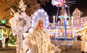 Frau in weißem, winterlichem Kostüm mit Zopf und großem, leuchtendem Stab auf einem festlich beleuchteten historischen Marktplatz mit Fachwerkhäusern.