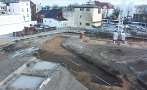 Baugrube mit archäologischen Ausgrabungen in einem ehemaligen Stadtgraben, umgeben von Wohnhäusern und einem Baukran.