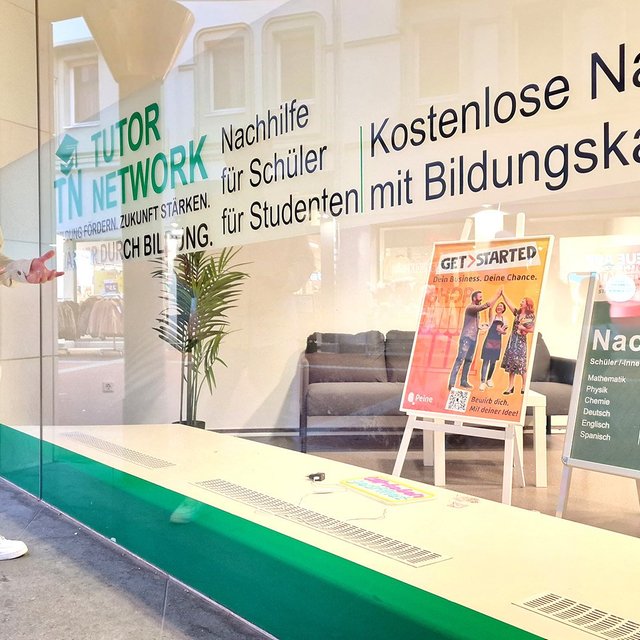 Ein Mann in weißer Kleidung steht vor einem Schaufenster mit der Aufschrift „Tutor Network“, hinter der Scheibe sind zwei Aufsteller mit Informationsplakaten zum Peiner Förderprojekt "GetStarted" und Nachhilfe zu sehen. 
