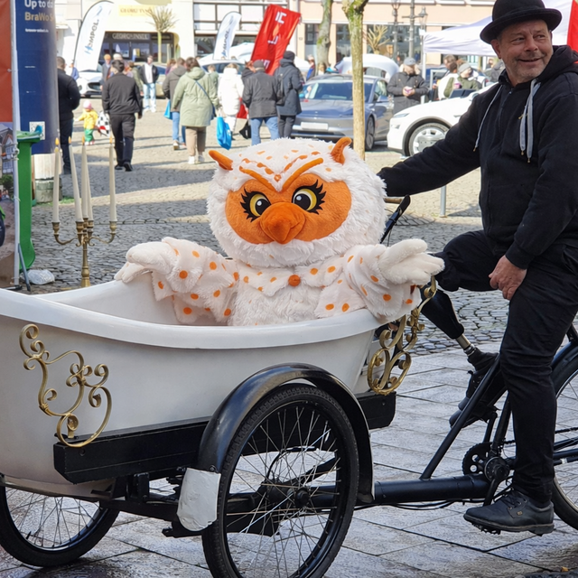 Uhlinchen sitzt lachend in einer weißen Badewanne auf einem Fahrrad-Anhänger beim BRAWO Mobility Spring in der Peiner Innenstadt, während ein Mann das Gefährt über den Historischen Marktplatz fährt.