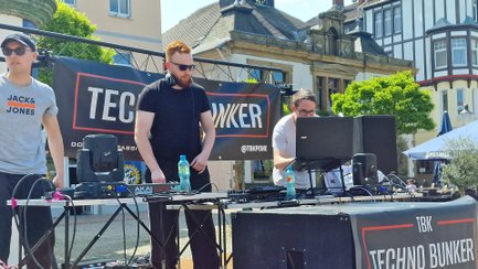 Mehrere Mitglieder des DJ-Kollektivs Techno Bunker Peine stehen an Mischpulten und Technik-Equipment bei einem Open-Air-Auftritt auf dem Historischen Marktplatz. Im Hintergrund hängt ein großes Banner mit dem Logo des Techno Bunker, während historische Gebäude und sonniges Wetter die Szene umrahmen.