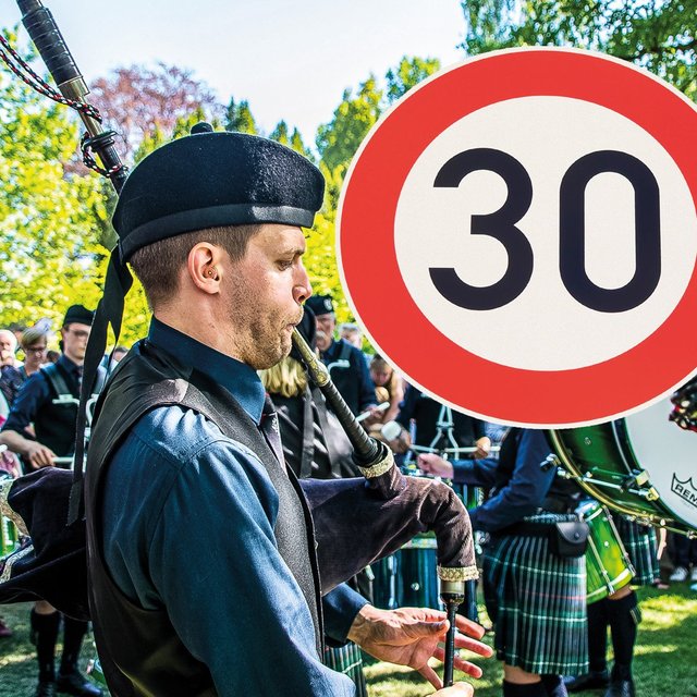 Ein Dudelsackspieler in traditioneller schottischer Kleidung spielt bei einem Highland Gathering, umgeben von einer Trommlergruppe und Zuschauern im Freien; im Vordergrund ist ein rundes Tempo-30-Verkehrsschild zu sehen.