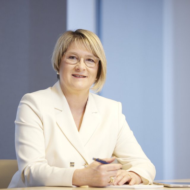 Frau mit kurzem blonden Haar in weißem Blazer sitzt an einem Tisch und hält einen Stift in der Hand.