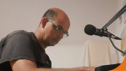 Musiker Christian Rebig sitzt an einem Klavier oder Keyboard und spielt konzentriert, während sich vor ihm ein Mikrofon auf einem Stativ befindet. Er trägt eine Brille und ein dunkles Shirt, der Hintergrund ist schlicht und hell gehalten.