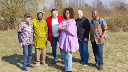 Die Band Pretty Cashanga steht mit sieben Personen auf einer Wiese und blickt in die Kamera. Einige tragen farbenfrohe Kleidung.
