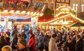 Belebter Weihnachtsmarkt mit festlich beleuchtetem Karussell, geschmückten Ständen und vielen Besuchern in Winterkleidung.
