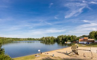 Badesee mit Sandstrand und Spielplatz an einem sonnigen Tag, Menschen entspannen am Ufer, Kinder spielen im Sand, im Hintergrund Bäume und ein Gebäude am Seeufer.